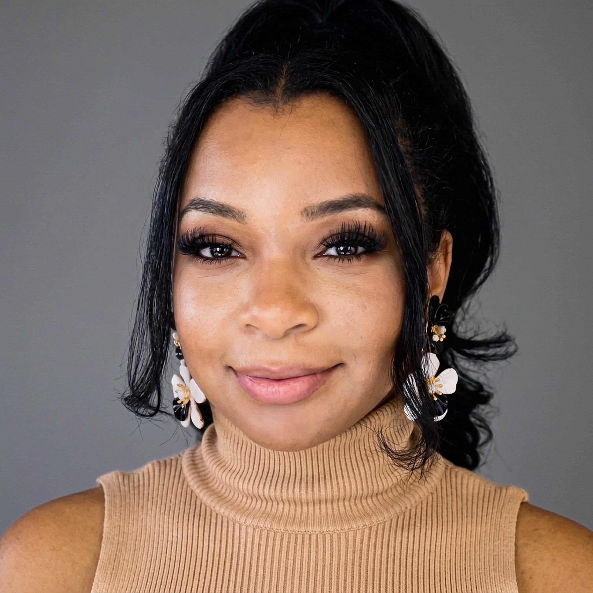 Portrait of Gillian Lyn Maxwell smiling, wearing a tan ribbed turtleneck and black-and-white floral earrings.